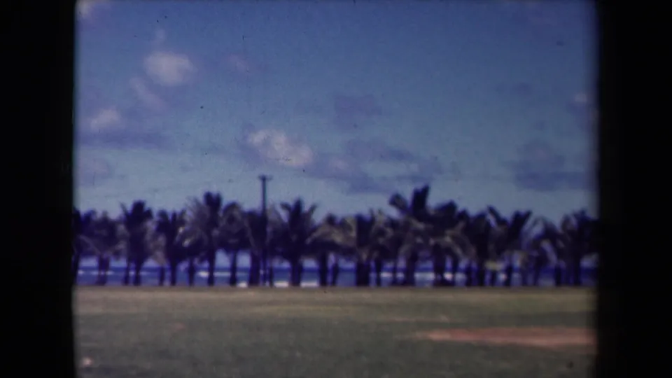 1944 Kauai 8mm frame with a soft coastal view of grass, palm trees, blue ocean, scattered clouds, and dark film edges.