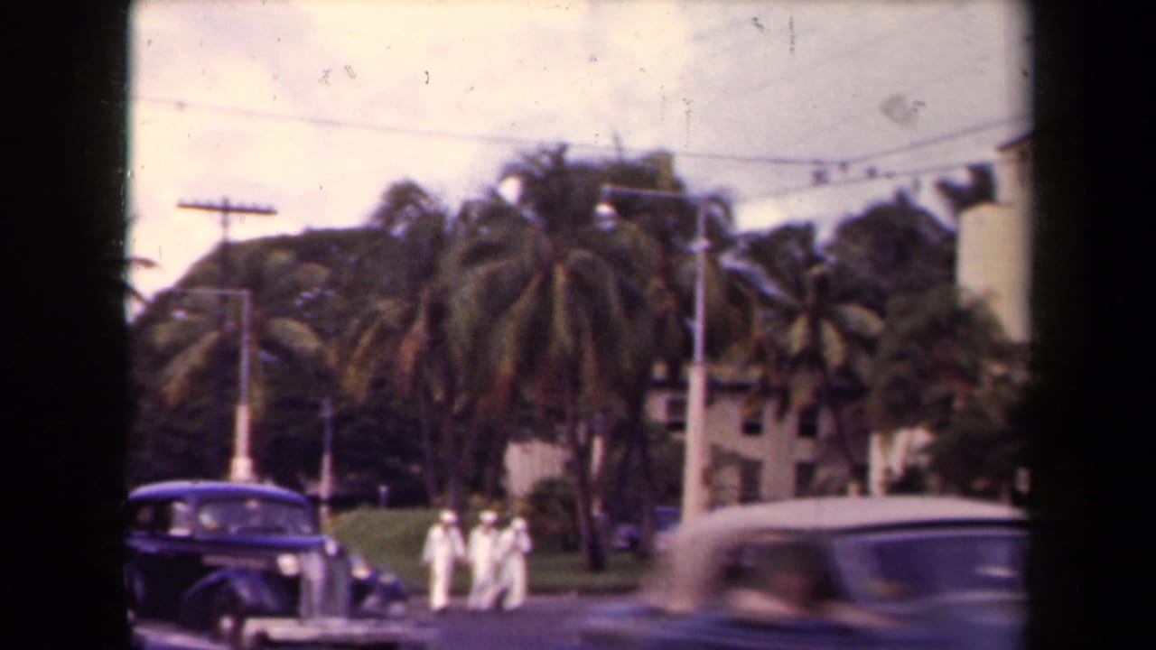 Three sailors in white uniforms walk along a palm-lined Hawaii street while dark 1940s-style cars move through the frame.