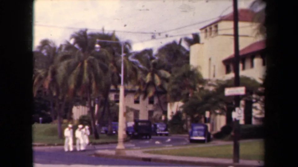Three sailors in white appear near a Hawaii street corner with palms, lawns, vintage cars, utility poles, and cream buildings in view.