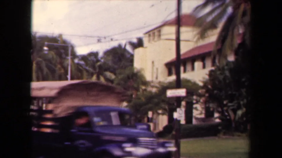 A dark 1940s-style car moves past the camera on a palm-lined Hawaii street with a cream building and street signs behind it.