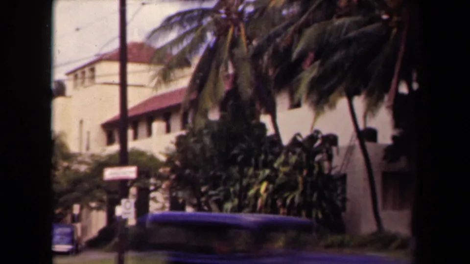 1944 Kauai home-movie frame showing palms, a stucco building with red roof, roadside signs, and the top of a car.