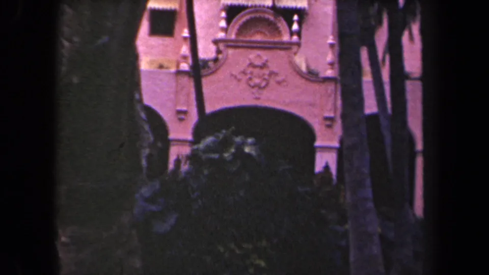 Palm-framed pink stucco facade with ornate arched entrance in 1940s Hawaii travel footage.