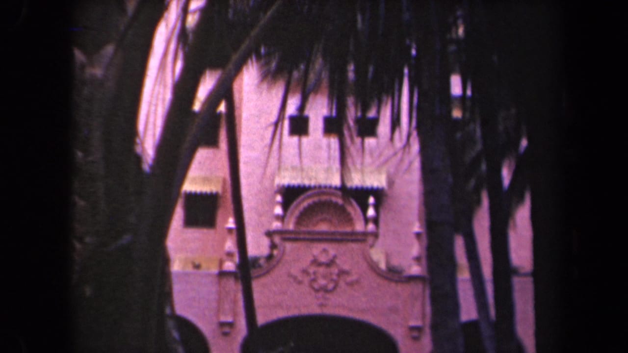 1944 Kauai home-movie frame showing a pink stucco resort entrance behind palm trunks and dark fronds.