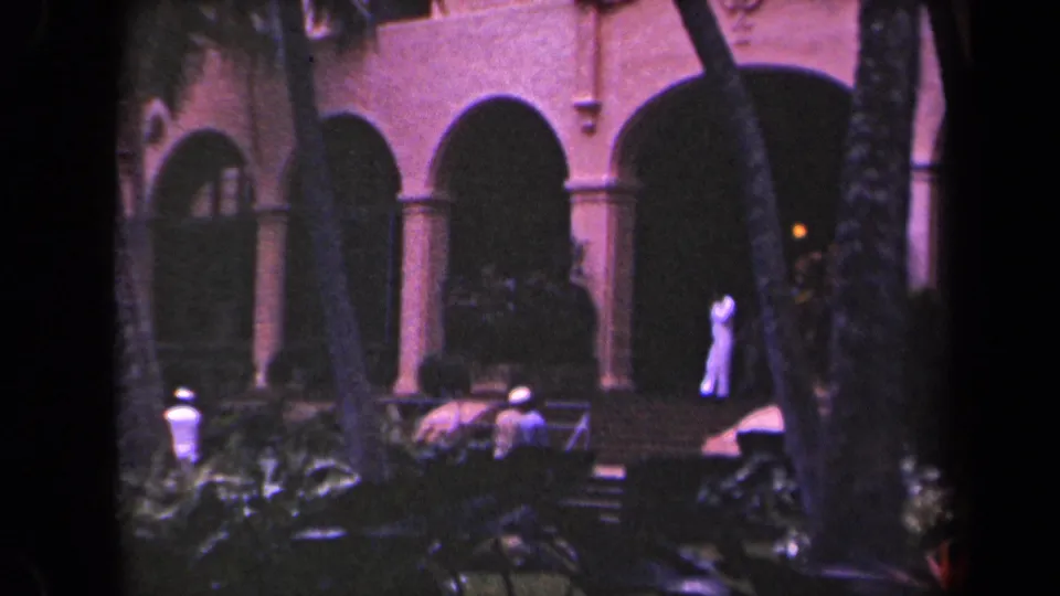 1944 Kauai resort courtyard with distant visitors, palm trunks, arched arcades, and a woman in light clothing near the entrance.