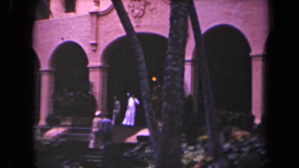 1944 Kauai home-movie frame showing visitors near steps and dark arches of a pink-toned historic-looking building.