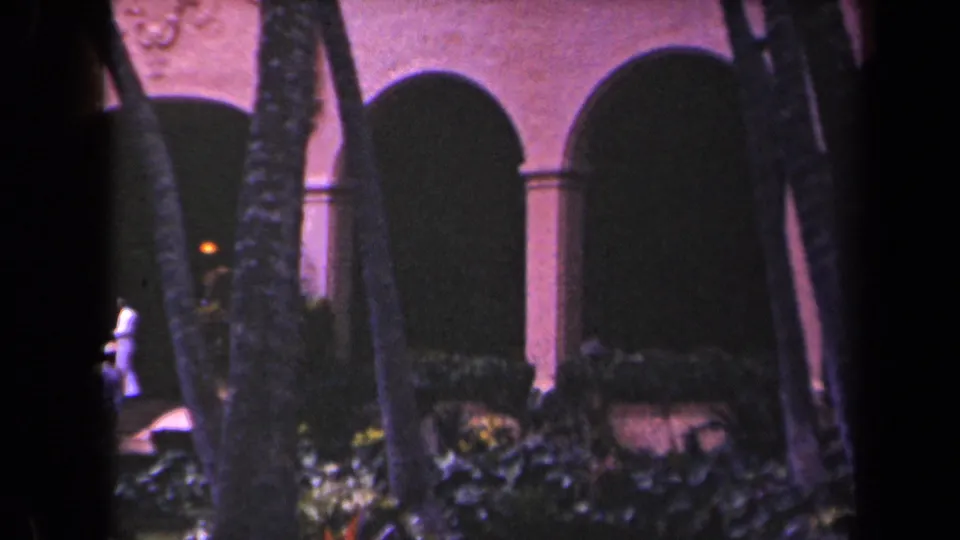 A visitor leaves the edge of the frame as the camera holds on palms, steps, and arched building openings in 1940s Kauai footage.