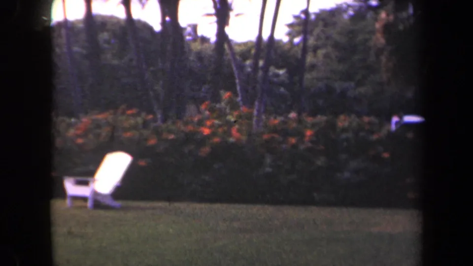 Final Kauai garden frame showing the white chair on the lawn, tropical plantings behind it, and a small pale object near the right edge.