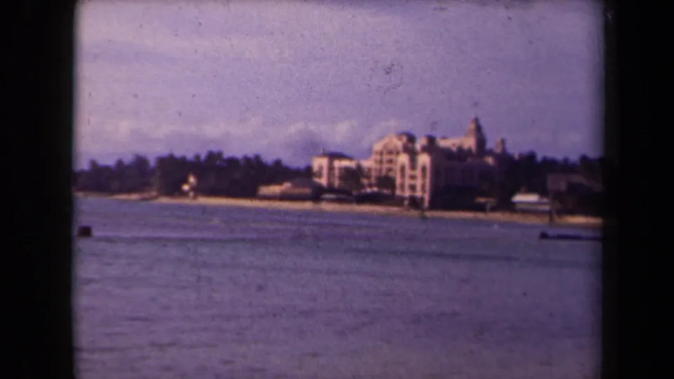 1940s 8mm view of Waikiki Beach shoreline, showing distant hotel buildings, calm water, sandy beach, and a hazy sky.