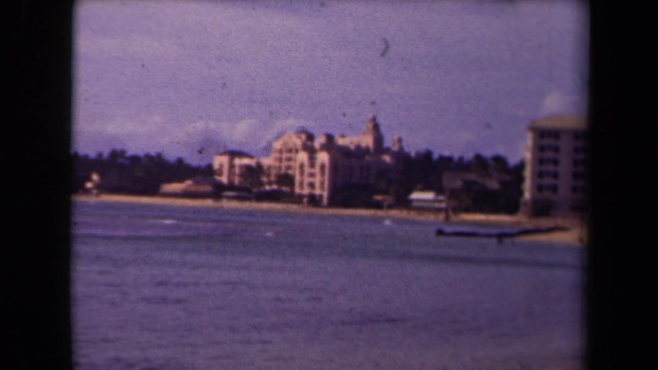 1944 Kauai home-movie frame showing calm water, sandy shoreline, distant trees, and large beachfront buildings under a hazy sky.