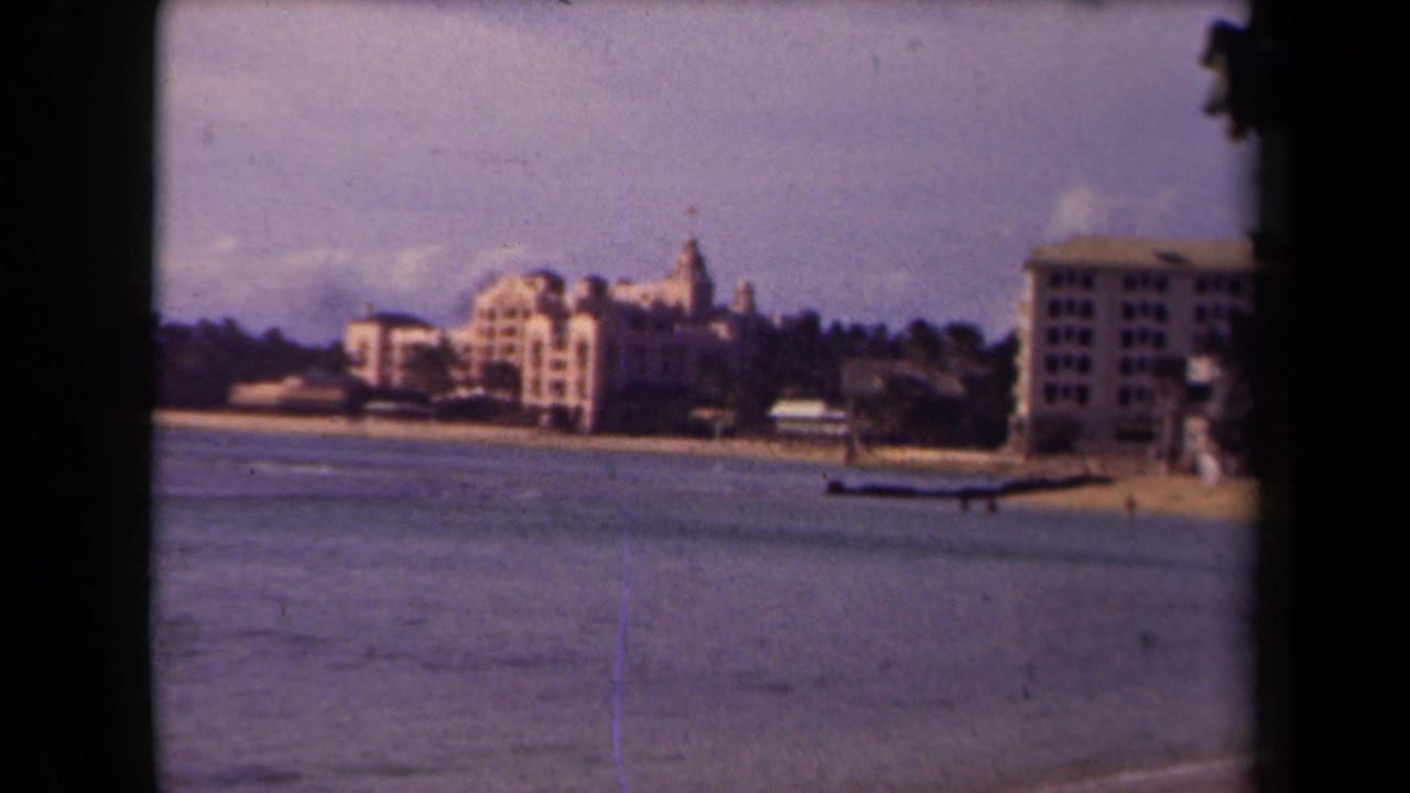 Beachfront buildings stand beyond calm water with a small shoreline structure at right.
