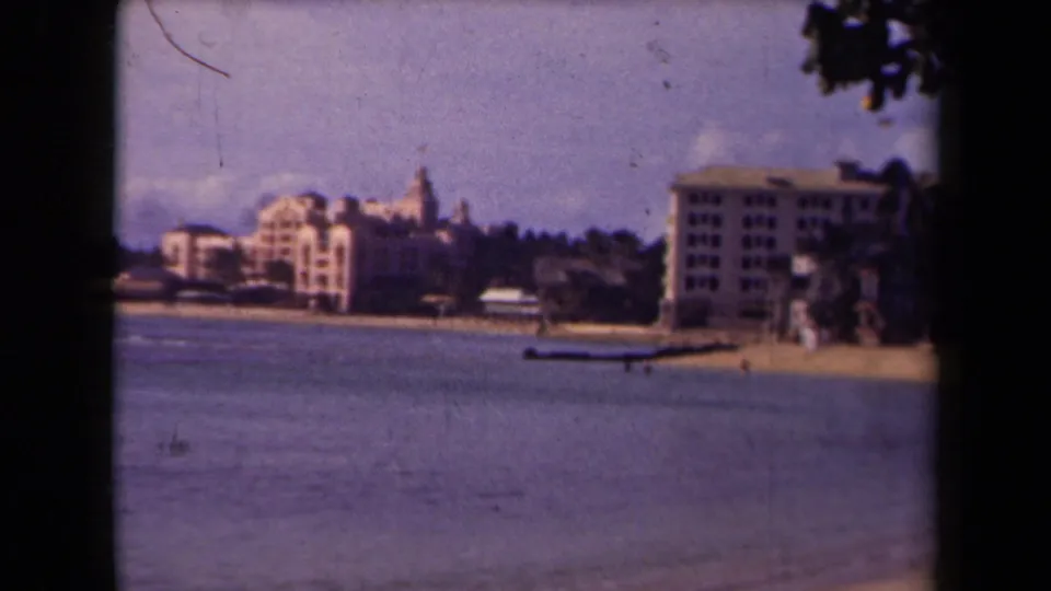 1944 Kauai home-movie frame showing a beachfront across calm water, with large buildings, trees, and dark foliage intruding at the right edge.