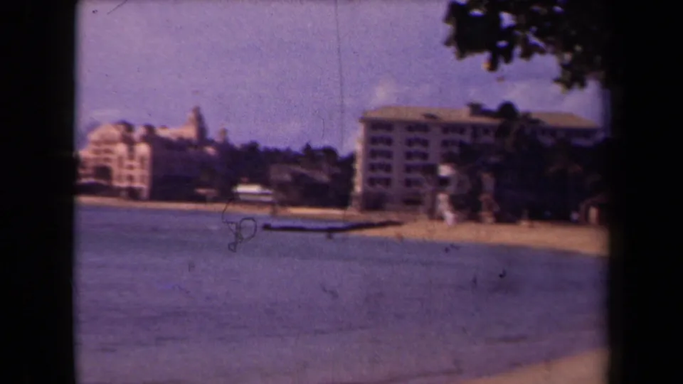 1944 Kauai frame showing calm water and an empty sandy shore with large buildings, distant trees, and dark leaves framing the upper right.