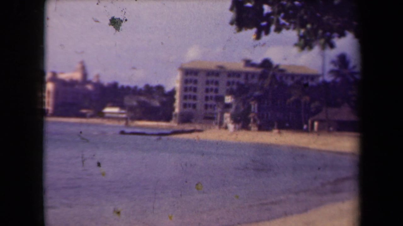 1944 Kauai 8mm frame showing a resort-like beachfront building, palms, sand, calm water, and a hazy tropical sky.