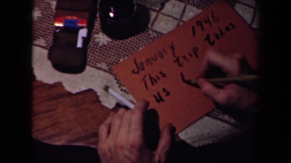 In France-related 1940s home-movie footage, adult hands write a January 1946 title card on a patterned tablecloth.