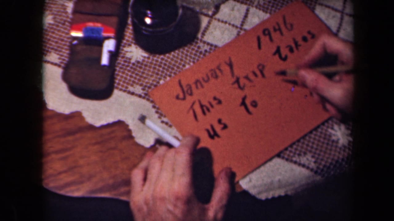 A hand continues lettering a January 1946 travel-title card, with tabletop objects and patterned cloth visible around it.