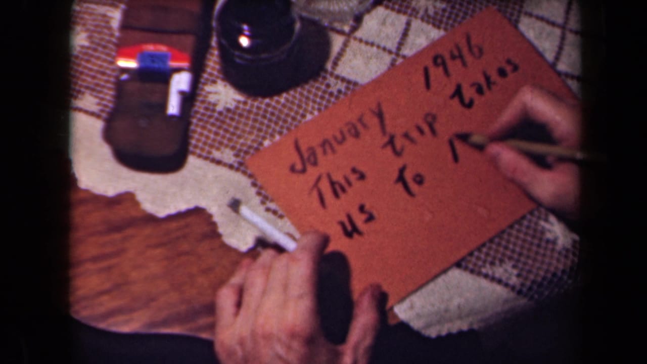 Hands write on a brown card resting on a patterned tablecloth beside a cigarette and dark glass object.