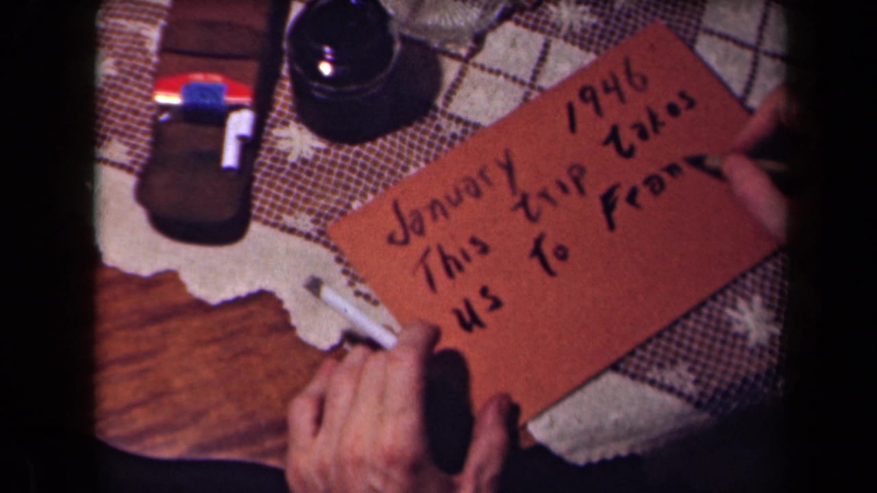 Close view of hands writing the phrase about a trip to France on a brown card, with a cigarette and glass object nearby.