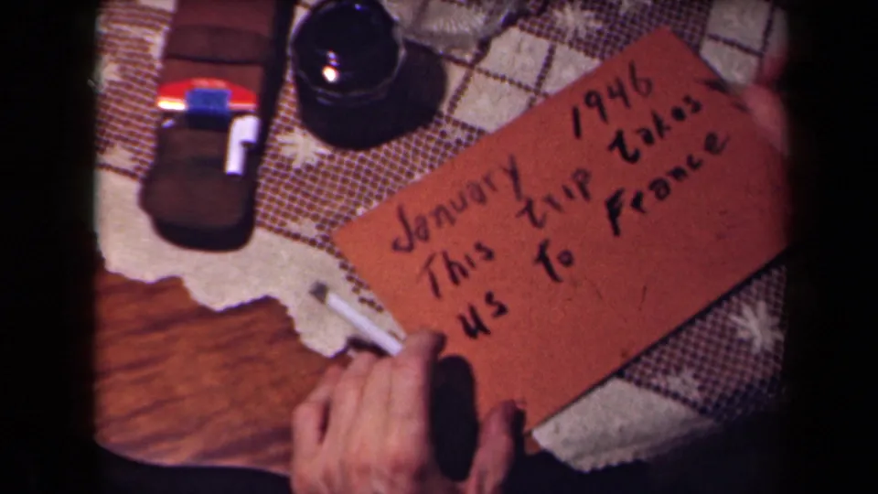 The brown card now clearly states a January 1946 trip to France, held by hands in a close 8mm tabletop shot.