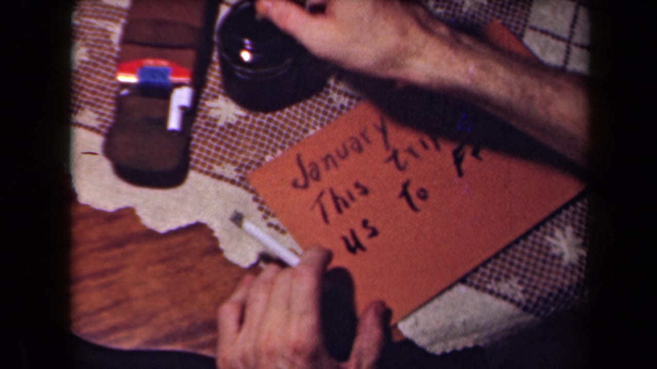 Close 1940s 8mm frame of hands writing travel text on a brown card on a domestic tabletop, with a cigarette nearby.