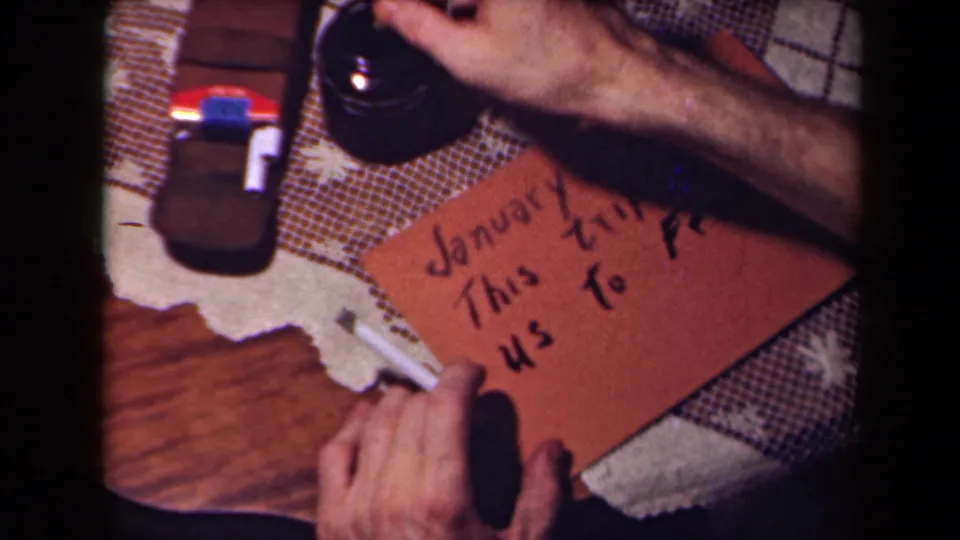 Close 1940s 8mm frame of hands writing travel text on a brown card on a domestic tabletop, with a cigarette nearby.
