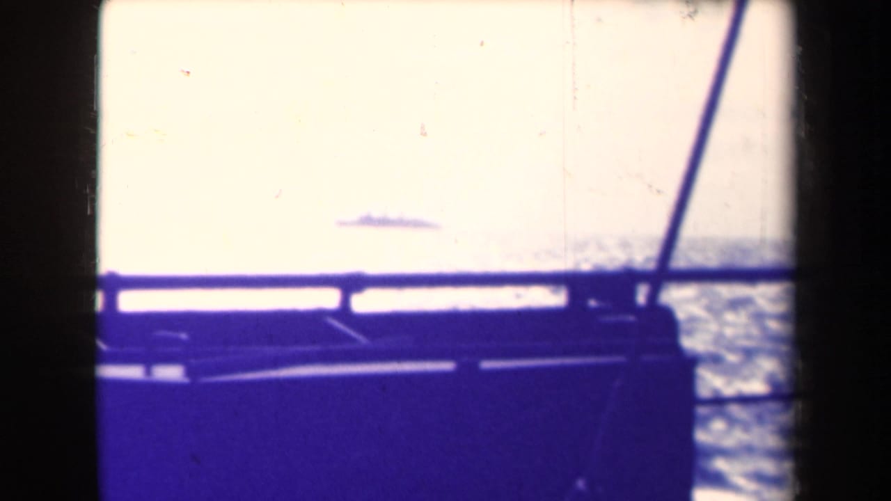 From a moving vessel, a small ship silhouette appears on the horizon beyond railing and window framing in 1945 home-movie footage.