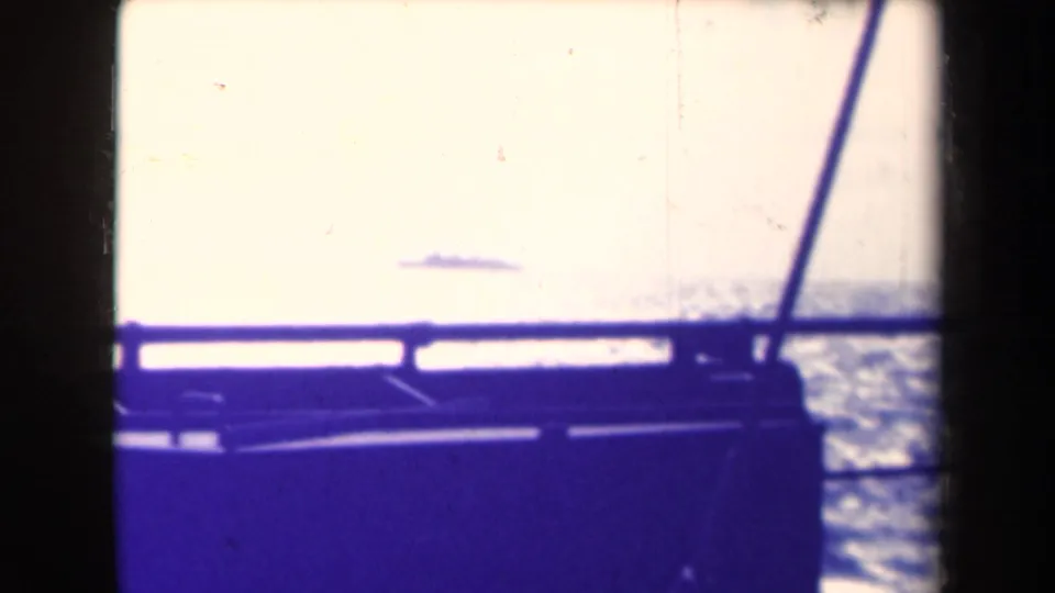 From a moving vessel, a small ship silhouette appears on the horizon beyond railing and window framing in 1945 home-movie footage.