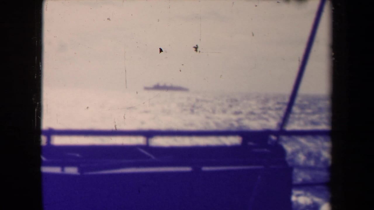 The distant Queen Mary appears as a small silhouette across open water, viewed through railing and a diagonal frame from another vessel.