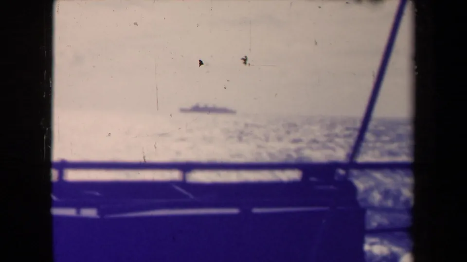 The distant Queen Mary appears as a small silhouette across open water, viewed through railing and a diagonal frame from another vessel.