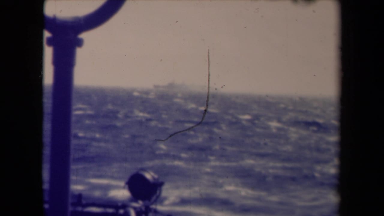Shipboard 8mm frame showing a faint distant vessel over choppy water, with dark rail structure and visible film wear.