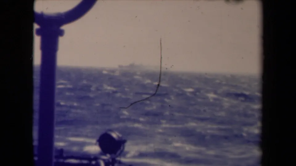 Shipboard 8mm frame showing a faint distant vessel over choppy water, with dark rail structure and visible film wear.