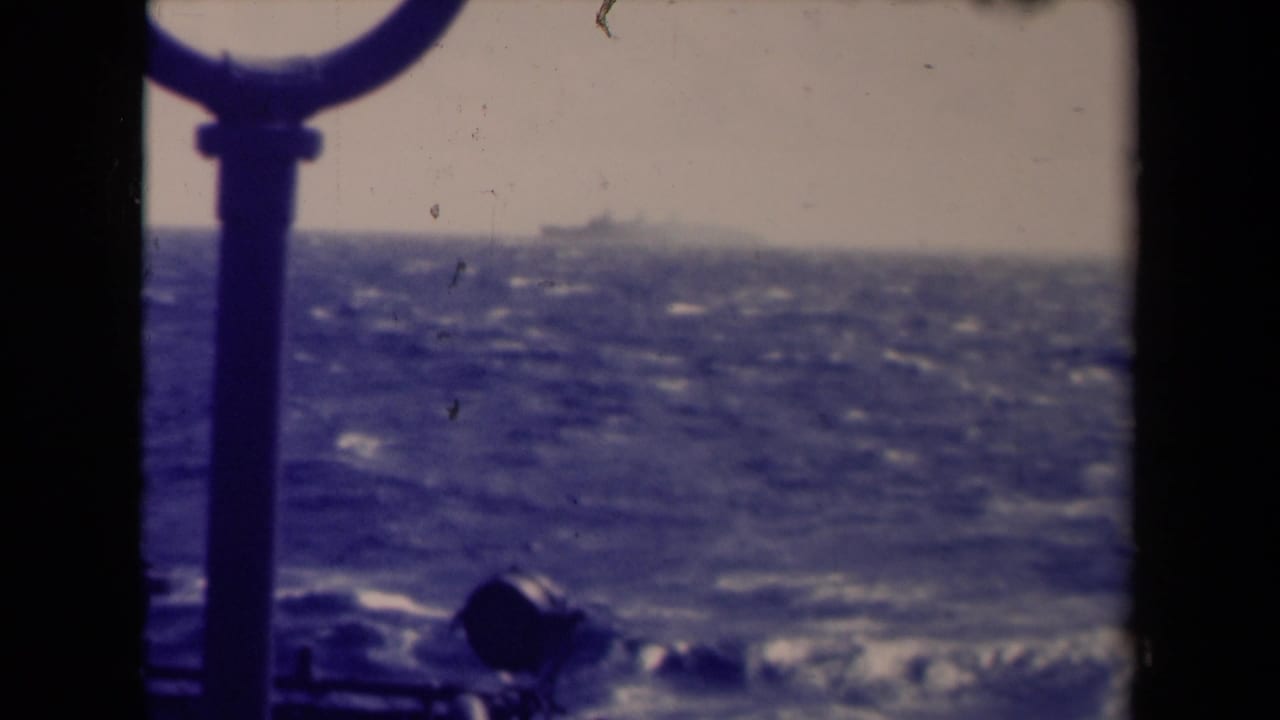 Rough sea seen from a ship deck with railing and a distant vessel on the hazy horizon.