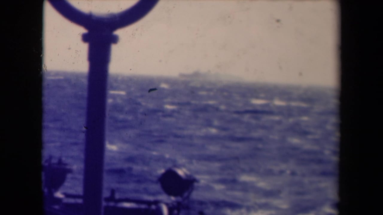 1940s 8mm maritime frame with a foreground ship fitting, choppy sea, and a faint distant vessel against a hazy horizon.