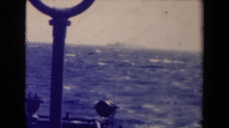 1940s 8mm maritime frame with a foreground ship fitting, choppy sea, and a faint distant vessel against a hazy horizon.