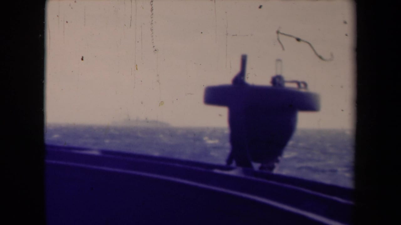 Shipboard 8mm frame from France metadata setting showing a dark deck structure, curved rail, rough water, and pale sky.