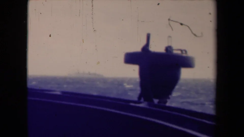 Shipboard 8mm frame from France metadata setting showing a dark deck structure, curved rail, rough water, and pale sky.