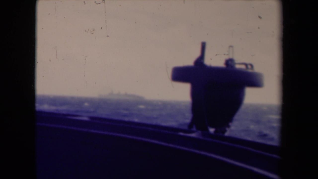 Shipboard 8mm frame with sloping deck rail, choppy blue water, and a dark foreground structure under a pale sky.