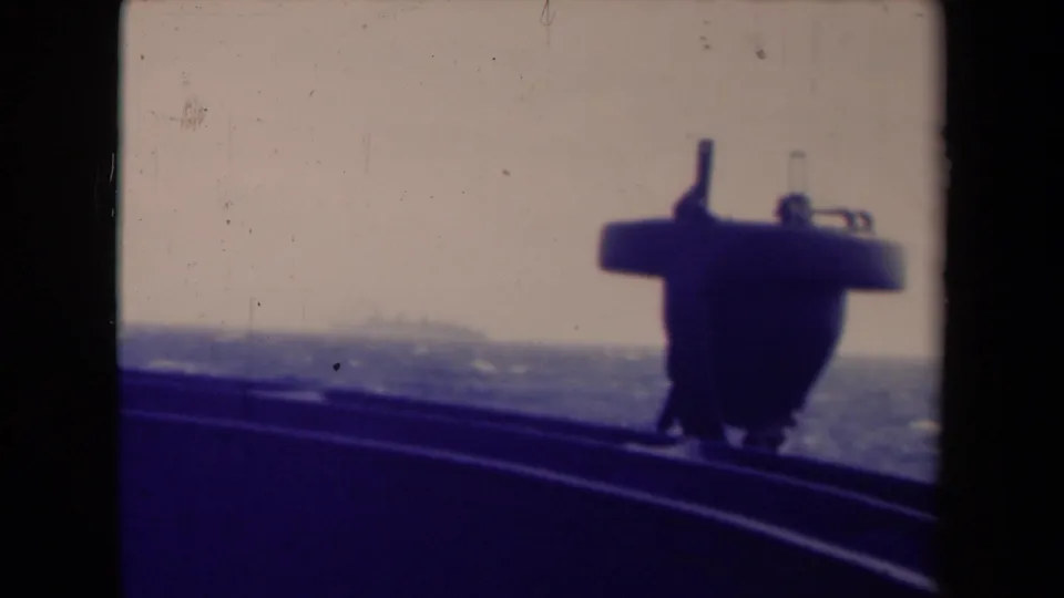 Final shipboard frame with a dark structure in front, choppy sea below, and a faint distant vessel or shoreline shape near the horizon.