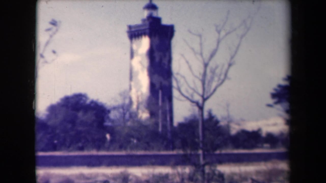 Tall lighthouse tower seen beyond bare trees and low greenery.