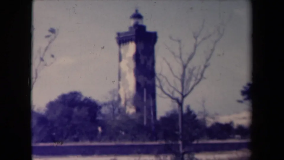 Color-tinted 8mm view of the Le Verdon lighthouse, with a stone tower, bare trees, and soft archival grain.