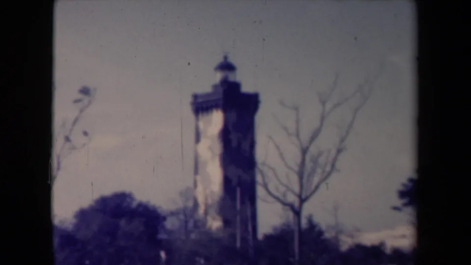 Le Verdon lighthouse remains centered in a grainy 1945 home-movie frame with bare branches and surrounding greenery.