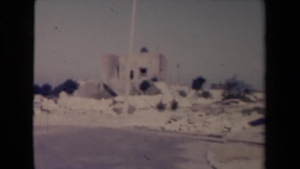 1945 France 8mm frame with a pale building, low roadside stones, bushes, and a curving road.