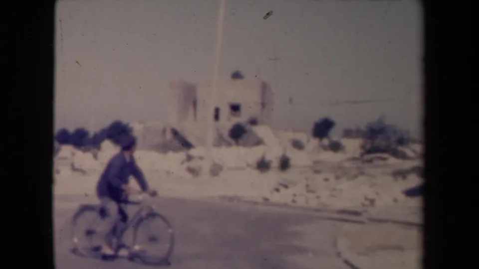 1945 France 8mm frame showing a man cycling along a road past a pale building and low roadside stones.