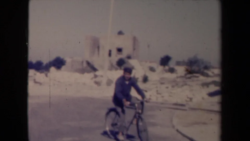 1945 France 8mm frame with a man on a bicycle near the camera, passing buildings and roadside stones.
