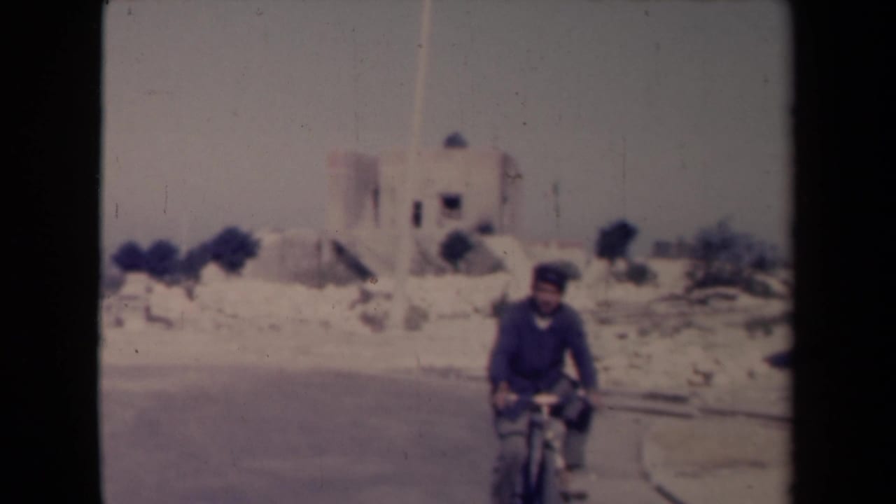 Man riding a bicycle on a dusty road near a pale church-like building.