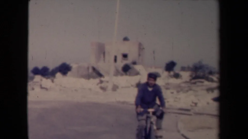 In 1940s France home-movie footage, a man rides a bicycle along a dusty road with a pale church-like building behind him.