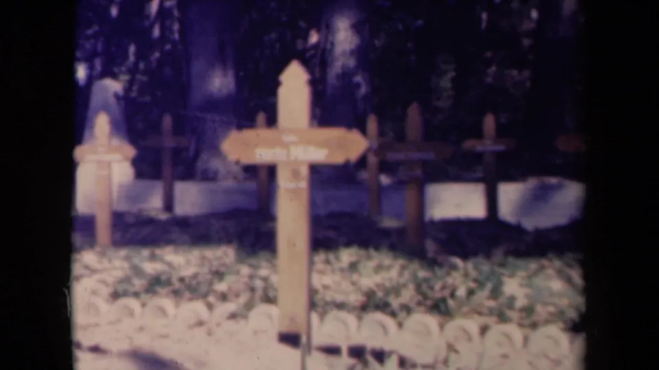 1945 France 8mm frame showing a wooden cemetery sign near crosses, grave markers, grass, border stones, and trees in soft daylight.