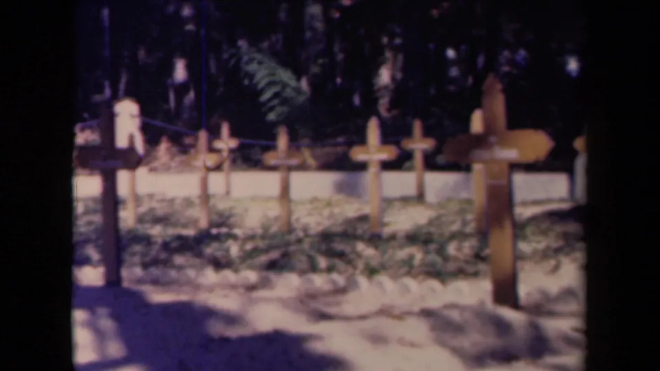 1940s France 8mm frame with a larger foreground wooden cross and several smaller grave markers set near a low white cemetery wall.