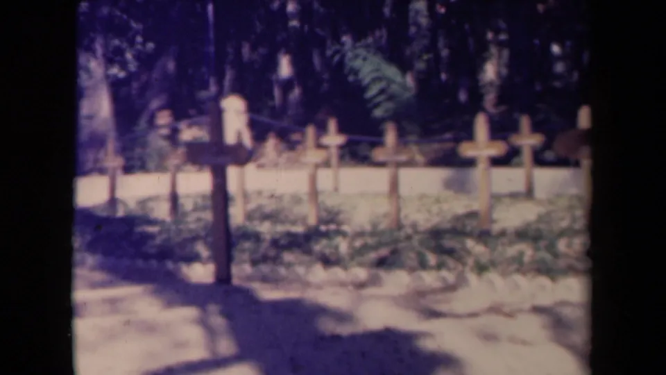 1940s France cemetery footage frame showing wooden cross grave markers across a shaded plot with pale soil and trees in the background.
