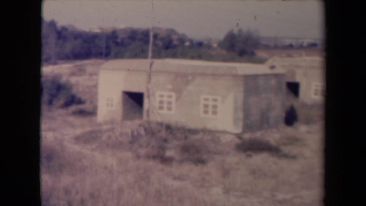 1945 France 8mm frame showing low utilitarian buildings in dry grass with small windows, open doorways, trees, and a pale horizon.