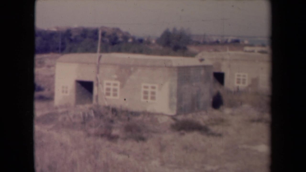 Weathered low structures viewed across dry grass with trees beyond.
